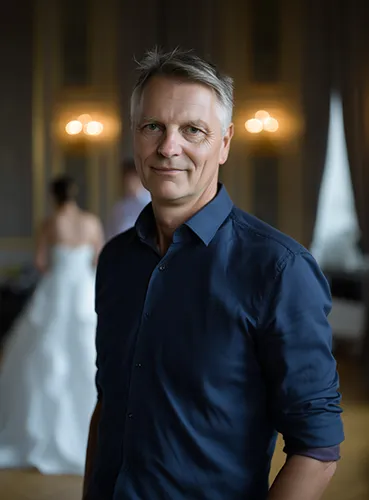 Ein Mann mittleren Alters mit grauen Haaren, der ein dunkelblaues Hemd trägt, steht in einem Raum und lächelt in die Kamera. Im unscharfen Hintergrund ist eine Person in einem weißen Kleid zu sehen, umgeben von sanftem Licht.