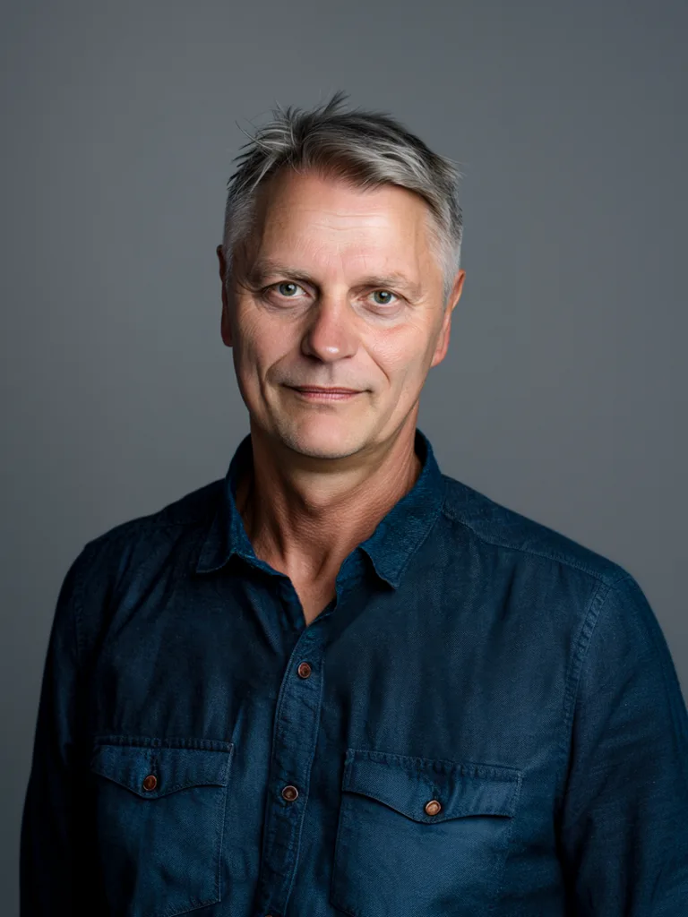 Axel von Onelovephoto Ein Mann mittleren Alters mit kurzen grauen Haaren, der ein dunkelblaues Hemd mit Knöpfen trägt, vor einem schlichten grauen Hintergrund steht und ruhig in die Kamera blickt – das ist Axel Rosenberg, Hochzeitsfotograf in Mannheim und Heidelberg.