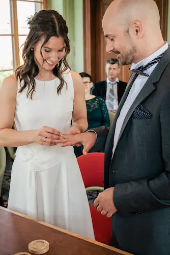Eine Frau in einem weißen Kleid steckt einem Mann während einer Zeremonie einen Ring an den Finger; beide lächeln. Im Hintergrund sitzen Menschen, die diesen Moment beobachten.