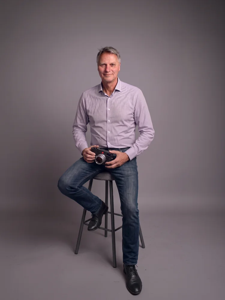 Ein Mann sitzt auf einem Hocker vor grauem Hintergrund; er trägt ein hellviolettes Hemd mit Knöpfen und Jeans. Er hält eine Vintage-Kamera in der Hand und lächelt in die Kamera – Axel von Onelove: Foto im Studio mit einer M-Leica, das den zeitlosen Geist der Fotografie einfängt.
