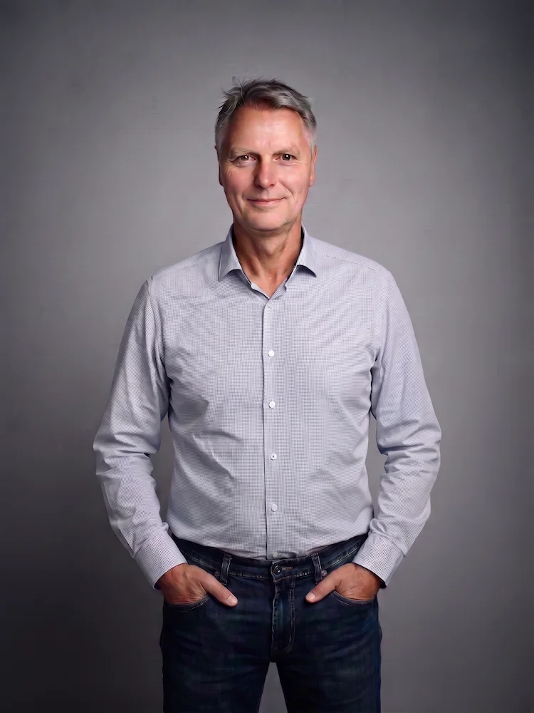 Axel von Onelovephoto Ein Mann mittleren Alters mit kurzen grauen Haaren steht vor einem grauen Hintergrund in Axel von Onelovephotos Studio. Er trägt ein hellgraues Hemd mit Knöpfen und eine dunkle Jeans, die Hände lässig in den Taschen und ein leichtes Lächeln auf den Lippen.