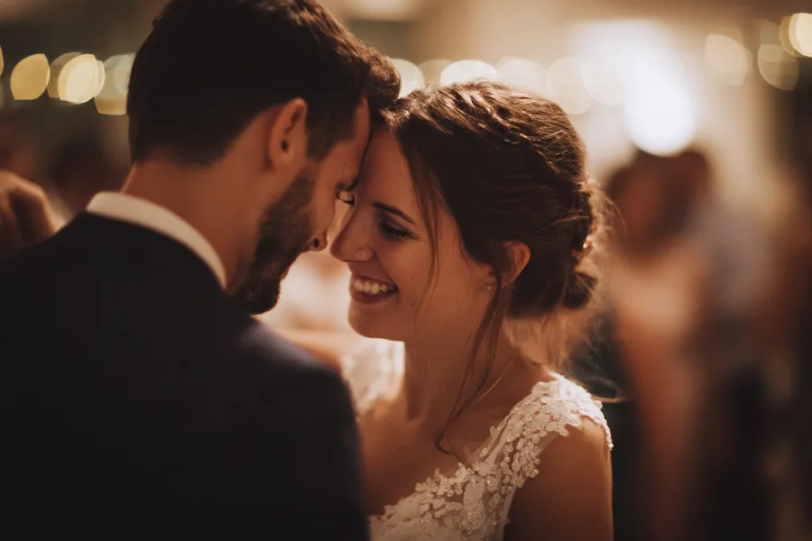 Ein Brautpaar lächelt sich liebevoll an, die Stirnen berühren sich, während sie auf ihrer Hochzeit zusammen tanzen. Warmes, sanftes Licht und verschwommene Gäste im Hintergrund schaffen eine romantische Atmosphäre.