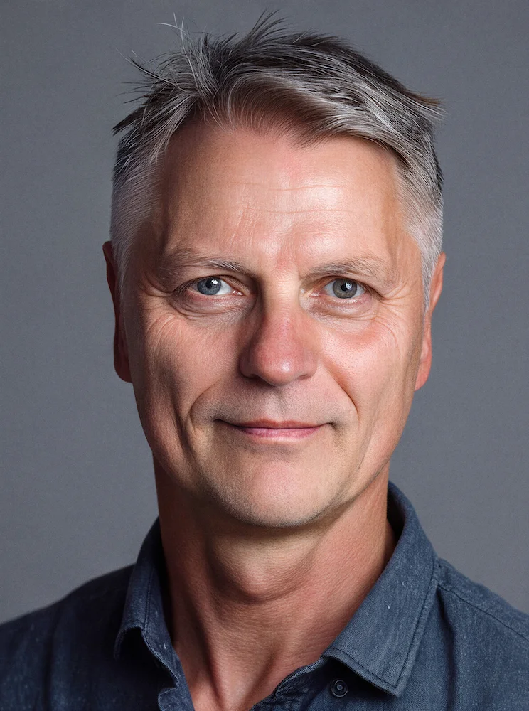 Ein Mann mittleren Alters mit kurzen grauen Haaren und blauen Augen, der ein blaues Hemd mit Knöpfen trägt, posiert vor einem schlichten grauen Hintergrund und blickt mit ausdruckslosem Gesicht direkt in die Kamera. Das ist Axel Rosenberg, ein renommierter Hochzeitsfotograf in Mannheim und Heidelberg.